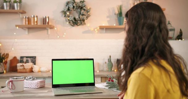Über die Schulter gesehen. Großaufnahme einer kaukasischen Frau in dekoriertem Haus, die auf den Bildschirm eines Laptops mit grünem Bildschirm in der Weihnachtsfestzeit blickt. Computer mit Chroma-Schlüssel. Technologiekonzept - Foto, Bild