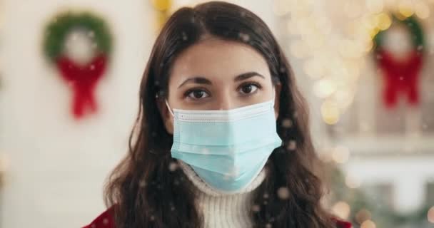 Közelkép portré vidám fiatal gyönyörű nő orvosi maszk áll a díszített karácsonyi utcában a városban, nézi a kamerát, és mosolyog újévkor. Kellemes ünnepeket a karantén koncepcióban - Felvétel, videó