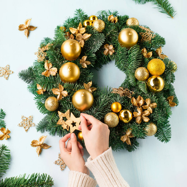 手は青い木製のテーブル、トップビューに出現素朴なクリスマスの花輪を作ります。現代のクリスマスは、青の背景に金のろうそく、枝や星で花輪を捧げます。冬休みの到来時間. - 写真・画像