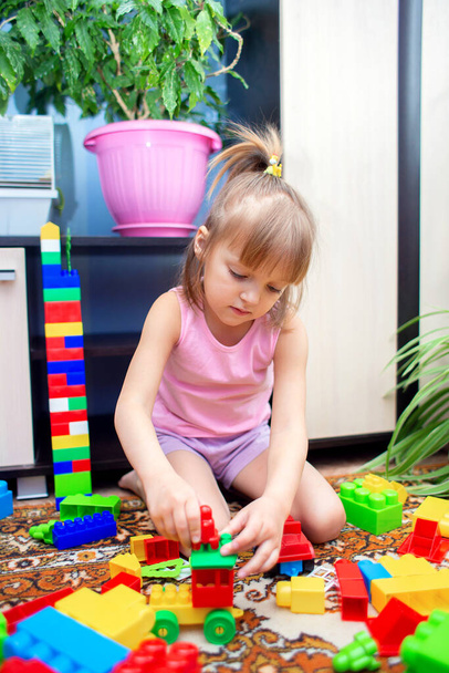 Ένα μικρό κορίτσι κάθεται στο πάτωμα στο σπίτι και παίζει ένα πολύχρωμο πλαστικό κατασκευαστή. Ένα παιδί παίζει με ένα τρενάκι - Φωτογραφία, εικόνα