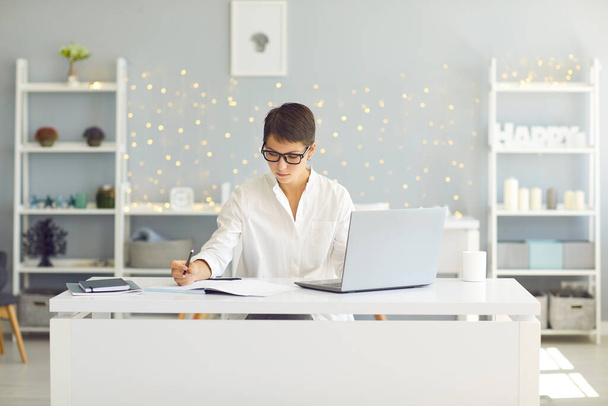Vážná mladá žena se zaměřením na práci nebo studium sedí u stolu s notebookem doma - Fotografie, Obrázek