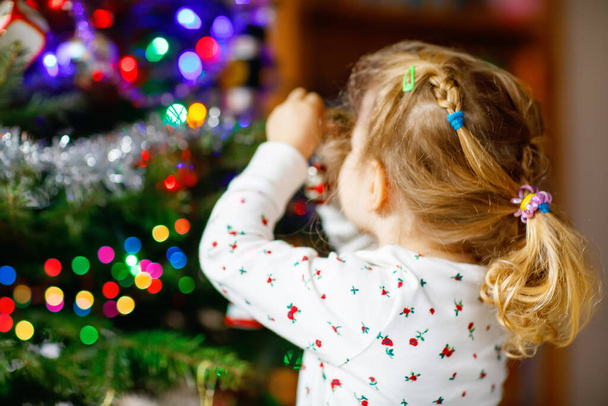 Urocza dziewczynka maluch w piżamie dekorowanie choinki z zabawką w uroczych rękach. Małe dziecko w koszuli nocnej stojące przy choince świątecznej. obchody tradycyjnego rodzinnego zimowego urlopu - Zdjęcie, obraz