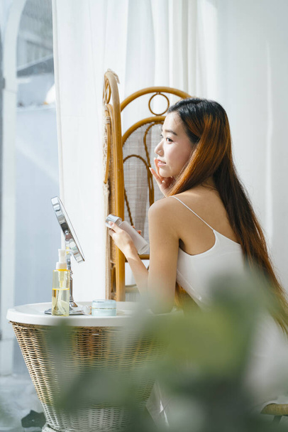 Side view - Jonge Aziatische vrouw doet make-up in de buurt van raam thuis. Natuurlijk licht. - Foto, afbeelding