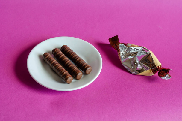 Three chocolates in white saucer on pink background. Sweet treats and candy wrapper on colored surface. Confectionery - Photo, Image