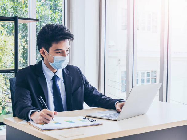 Junger hübscher asiatischer Geschäftsmann im Anzug trägt eine schützende Gesichtsmaske, arbeitet am Laptop und schreibt und überprüft den Businessplan auf dem Diagramm, während er im Büro in der Nähe eines riesigen Glasfensters sitzt. - Foto, Bild