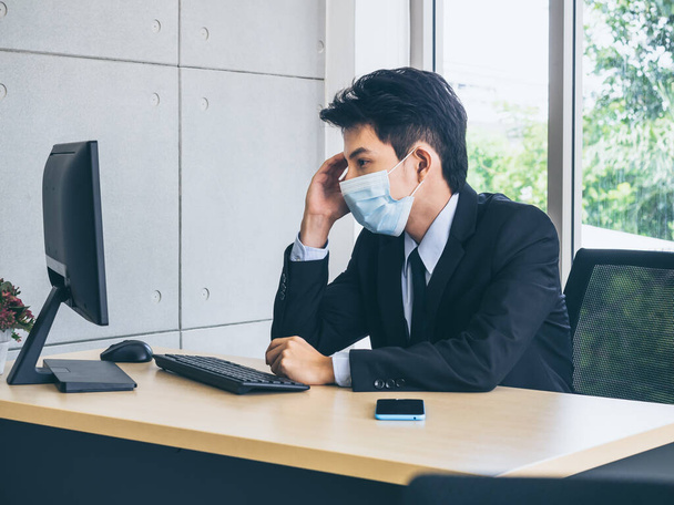 Jonge Aziatische zakenman in pak dragen beschermende gezichtsmasker werken op de computer en denken met spanning met moe gezicht met smartphone op kantoor in de buurt van enorme glazen raam. - Foto, afbeelding