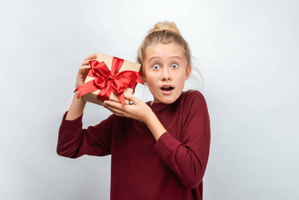 Charming blonde girl 12-15 years old with gift decorated with ribbon posing with surprised face expression. Studio shot white background, isolated. New Year, Women's Day, Birthday, Holiday concept - Photo, Image