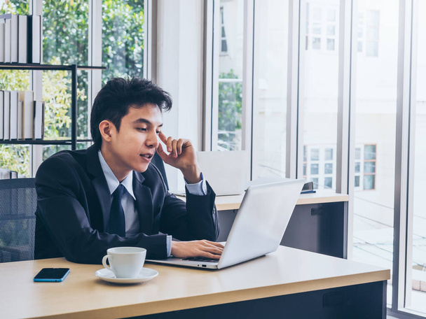 Młody azjatycki biznesmen w garniturze pracujący na laptopie i myślący ze zmęczoną twarzą ze smartfonem i filiżanką kawy na biurku w biurze w pobliżu szklanego okna z przestrzenią do kopiowania. - Zdjęcie, obraz