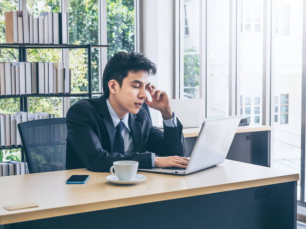 Молодой азиатский бизнесмен в костюме работает за ноутбуком и думает с напряжением с уставшим лицом со смартфоном и кофейной чашкой на столе в офисе возле стеклянного окна. - Фото, изображение
