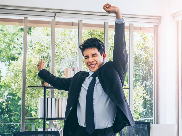 Young handsome Asian businessman in suit standing and stretching exercise near desk in office near huge glass window with sunlight. - Foto, imagen