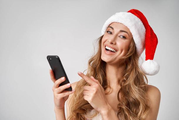 Woman christmas Santa Hat white studio background with smartphone in hand Beautiful caucasian female curly hair portrait. Happy person showing finger cell phone Holiday concept Teeth smiling  - Fotó, kép