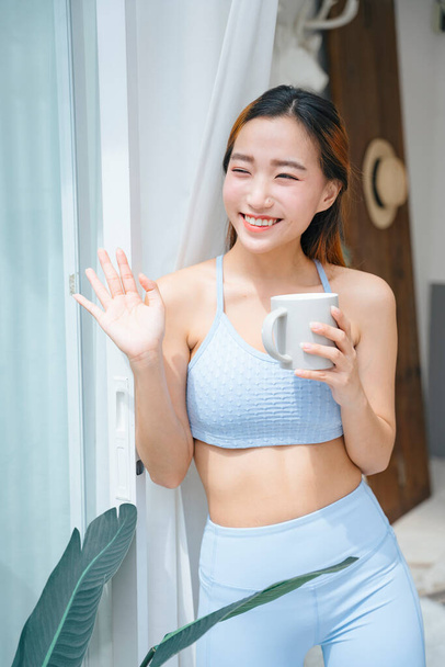 Beautiful sporty asian woman wearing sportbra holding cup of coffee at home balcony. - Photo, Image