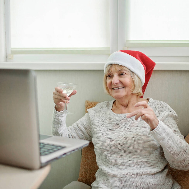 Celebración segura en línea. Mujer mayor sosteniendo una copa de vino y celebrando la Navidad con su familia virtualmente a través de Internet y portátil en casa. Videollamada. Vacaciones lejanas, estilo de vida interior - Foto, Imagen