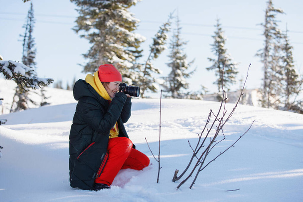 Női fotós a sífelszerelés készít képeket ül a hóban. Egy kamerát tart hatalmas lencsével. Téli ruházat: nadrág és meleg kabát, kötött sapka, síszemüveg. Szelektív fókusz. - Fotó, kép
