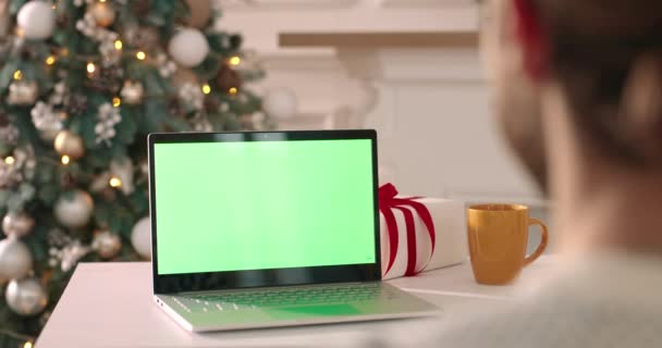 Přes rameno pohled na mladého muže sedí ve zdobené místnosti a dívá se na notebook se zelenou obrazovkou na Vánoce. Běloch na Štědrý den pomocí počítače s klíčem chroma. Zavřít. Technický koncept - Záběry, video