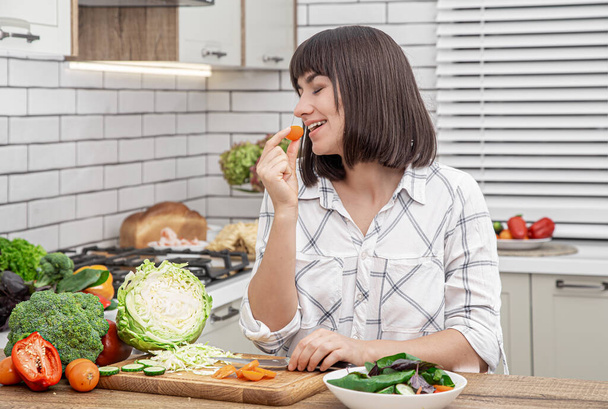 Beautiful brunette smiles and cuts vegetables on a salad on the background of a modern kitchen interior. - Photo, Image