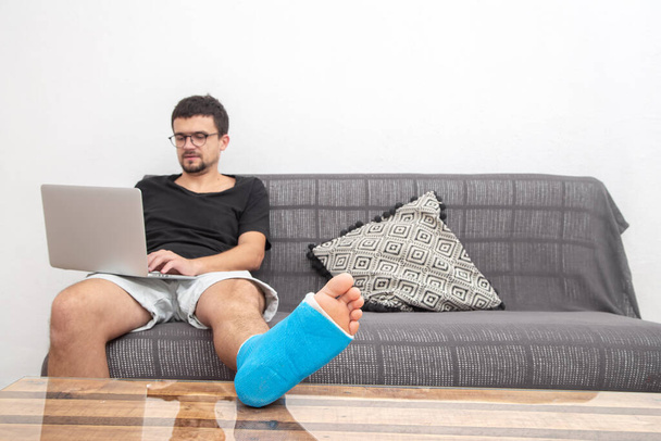 Mann mit Brille mit gebrochenem Bein in blauer Schiene zur Behandlung von Verletzungen durch Verstauchung am Laptop auf der heimischen Couch. - Foto, Bild