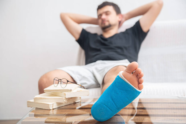 Mężczyzna ze złamaną nogą w niebieskiej szynie do leczenia urazów po skręceniu kostki czytanie książek w domu rehabilitacji. - Zdjęcie, obraz