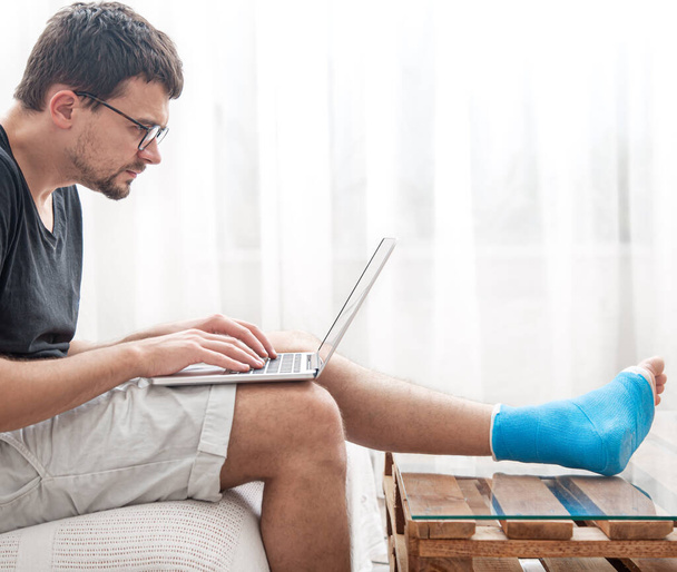 Mladý muž se zlomenou nohou v modré dlaze pro léčbu poranění a vymknutí kotníku používá doma notebook. - Fotografie, Obrázek