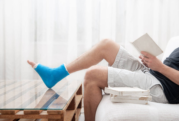 Ένας άντρας με σπασμένο πόδι σε γύψο διαβάζει βιβλία με φόντο το εσωτερικό του δωματίου.. - Φωτογραφία, εικόνα