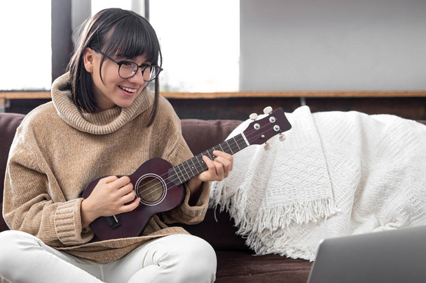 Młoda stylowa dziewczyna w okularach uczy się grać na ukulele. Koncepcja edukacji internetowej, wychowania domowego. - Zdjęcie, obraz