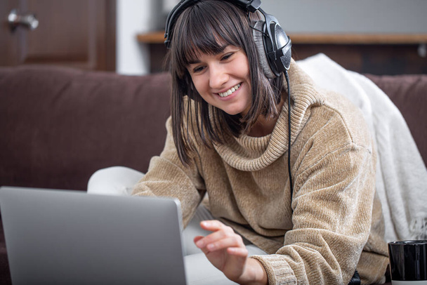Młoda piękna brunetka uśmiechnięta w przytulnym swetrze i patrząca na ekran laptopa. Koncepcja szkolenia online, pracy zdalnej, komunikacji wideo. - Zdjęcie, obraz