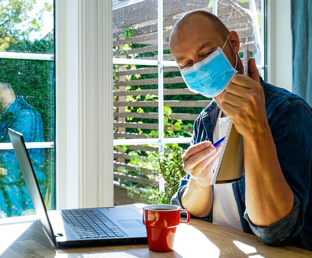 Ένας ενήλικος άνδρας που φοράει μάσκα προσώπου, εργάζεται από το σπίτι και κάθεται μπροστά σε ένα φορητό υπολογιστή δείχνοντας χαρτιά κατά τη διάρκεια μιας τηλεδιάσκεψης. . Υψηλής ποιότητας φωτογραφία - Φωτογραφία, εικόνα