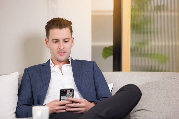 Деловой человек с чемоданом с помощью приложения для смартфона сидит на диване - Фото, изображение