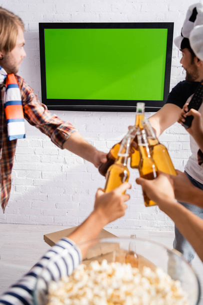 fanáticos del fútbol multiétnico tintinean botellas de cerveza mientras ven el campeonato en la televisión cerca de palomitas de maíz en un fondo borroso - Foto, Imagen
