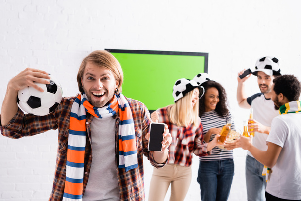взволнованный человек, держащий футбольный мяч и смартфон с пустым экраном рядом с многонациональными друзьями на размытом фоне - Фото, изображение