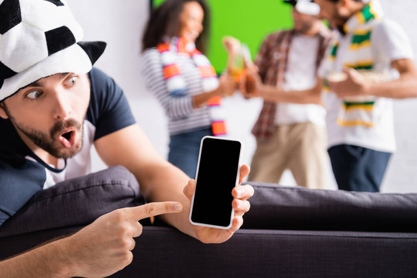 šokovaný muž ukazuje prstem na smartphone s prázdnou obrazovkou v blízkosti multietnické přátele na rozmazaném pozadí - Fotografie, Obrázek