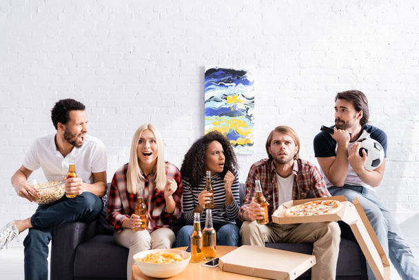 обеспокоенные и взволнованные футбольные болельщики смотрят чемпионат с пивом, пиццей и чипсами дома - Фото, изображение