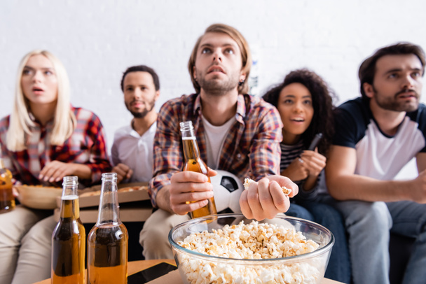концентрированный человек, принимающий попкорн во время просмотра футбольных соревнований рядом с многонациональными друзьями на размытом фоне  - Фото, изображение