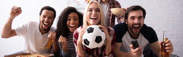 cheerful woman holding soccer ball while watching championship with excited multiethnic friends, banner - Photo, Image