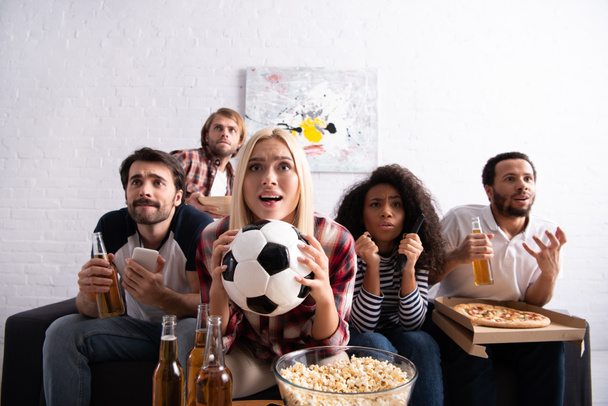 worried woman holding soccer ball while watching football championship with tense multiethnic friends - Foto, afbeelding