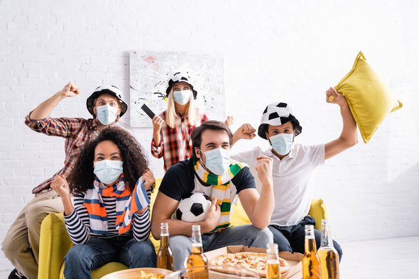 excited multiethnic friends in medical masks and football fans hats watching championship at home - Фото, зображення