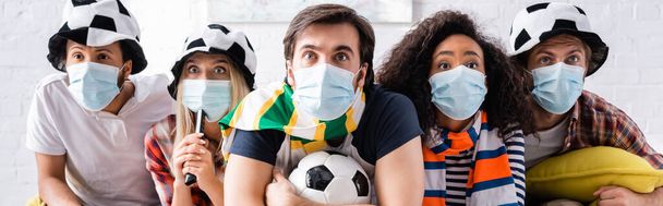 ανήσυχοι πολυεθνικοί φίλοι σε ποδοσφαιρικούς οπαδούς καπέλα και ιατρικές μάσκες βλέποντας πρωτάθλημα στο σπίτι, πανό - Φωτογραφία, εικόνα