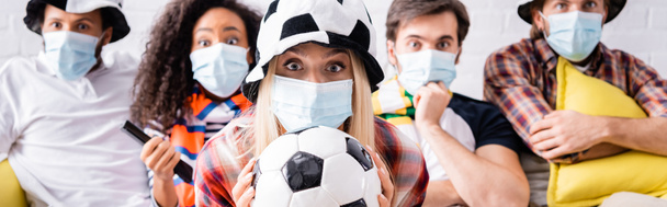 woman in football fan hat holding soccer ball near shocked multicultural friends in medical masks on blurred background, banner - Photo, Image