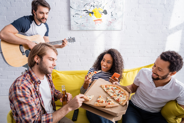 multicultural friends eating pizza on sofa while young man playing acoustic guitar - Foto, Bild