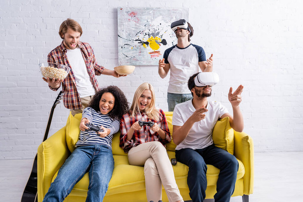 KYIV, UKRAINE - OCTOBER 19, 2020: cheerful multicultural friends using vr headsets and gaming with joysticks during party - Foto, afbeelding