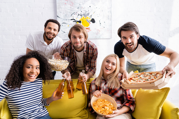 cheerful multicultural friend with beer, pizza and snacks looking at camera during party - 写真・画像