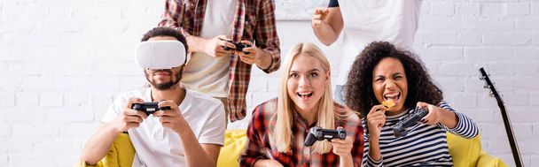 KYIV, UKRAINE - OCTOBER 19, 2020: cheerful african american woman eating chips while playing video game with multicultural friends - Φωτογραφία, εικόνα