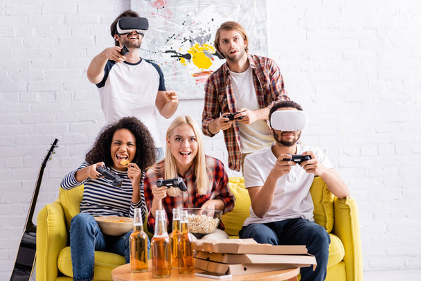 KYIV, UKRAINE - OCTOBER 19, 2020: cheerful multiethnic friends using vr headsets and playing video game during party - Фото, изображение