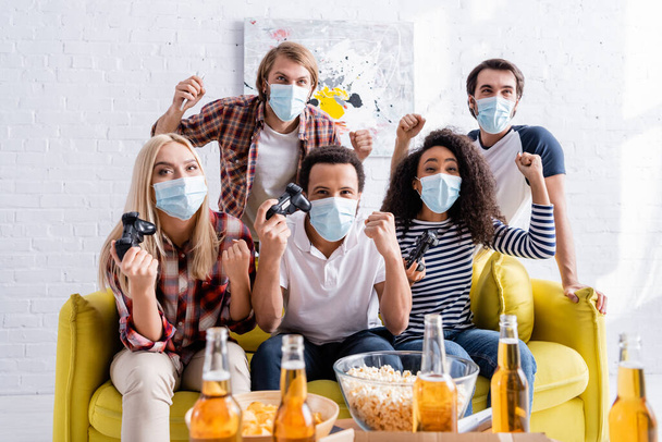 KYIV, UKRAINE - OCTOBER 19, 2020: excited multiethnic friends in medical masks showing win gesture while playing video game near beer on blurred foreground - Foto, Bild
