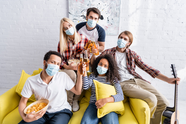 multiethnic přátelé v lékařských maskách při pohledu na kameru při cinkání lahví piva během párty - Fotografie, Obrázek
