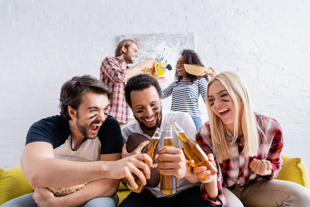hombre emocionado gritando mientras tintinea botellas de cerveza con amigos multiculturales con caras pintadas - Foto, Imagen