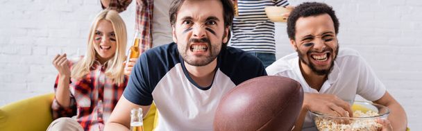 young man holding rugby ball and grimacing while watching championship near multiethnic friends, banner - Foto, Imagem