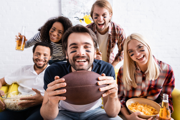 excited man holding rugby ball while watching championship near multiethnic friends with painted faces on blurred background - Photo, Image