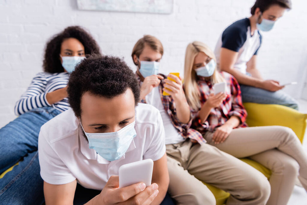 Afrikaans amerikaanse man in medische maskers chatten op mobiele telefoon in de buurt van multiculturele vrienden op wazige achtergrond - Foto, afbeelding