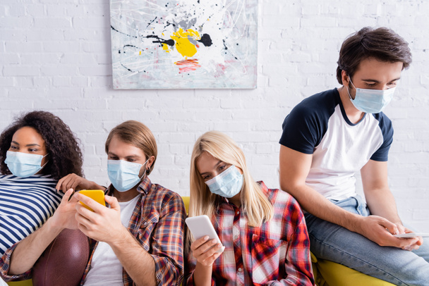 többnemzetiségű barátok orvosi maszkok üzenetküldés mobiltelefonok, miközben ül a kanapén - Fotó, kép